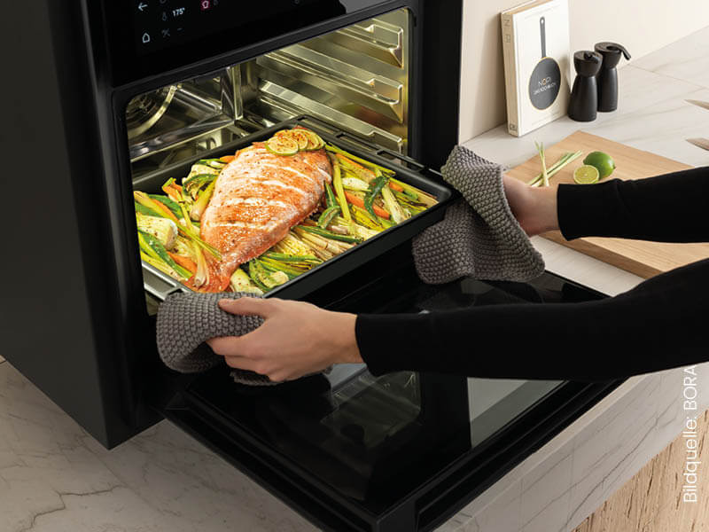 Frau nimmt Blech mit Fisch und Gemüse aus Dampfgarbackofen