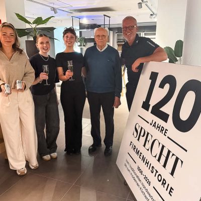 Mitarbeiter und Familie stoßen mit Getränken bei der Jubiläumsfeier SPECHT im modernen Küchenstudio vor, neben einem Schild „120 Jahre SPECHT Firmengeschichte“.