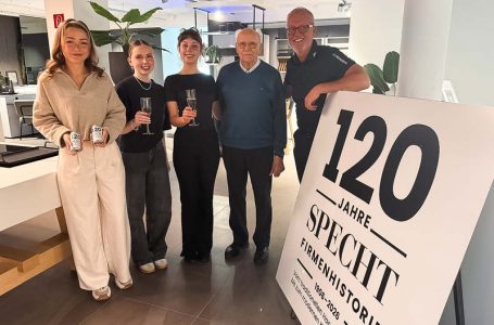 Mitarbeiter und Familie stoßen mit Getränken bei der Jubiläumsfeier SPECHT im modernen Küchenstudio vor, neben einem Schild „120 Jahre SPECHT Firmengeschichte“.