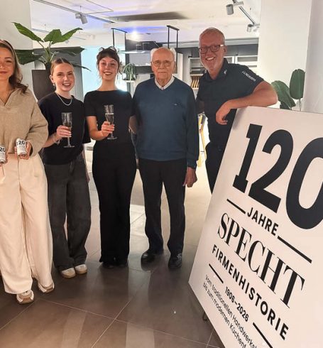 Mitarbeiter und Familie stoßen mit Getränken bei der Jubiläumsfeier SPECHT im modernen Küchenstudio vor, neben einem Schild „120 Jahre SPECHT Firmengeschichte“.