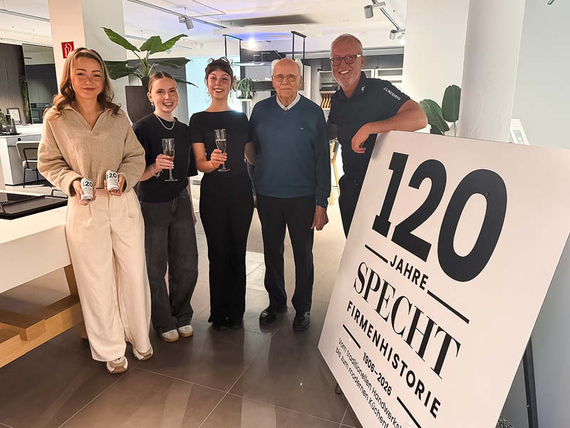 Mitarbeiter und Familie stoßen mit Getränken bei der Jubiläumsfeier SPECHT im modernen Küchenstudio vor, neben einem Schild „120 Jahre SPECHT Firmengeschichte“.