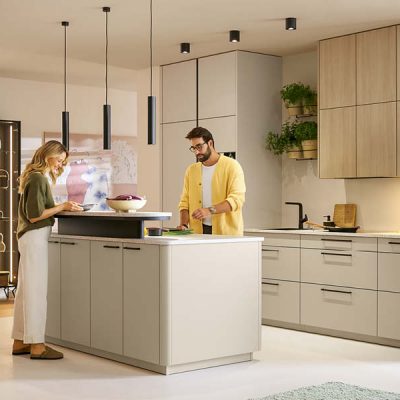 Moderne Schüller Küche mit Kochinsel in Beige und Holzoptik, integriertem Stauraum und Hängelampen, zwei Personen bereiten gemeinsam Speisen zu