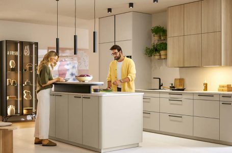 Moderne Schüller Küche mit Kochinsel in Beige und Holzoptik, integriertem Stauraum und Hängelampen, zwei Personen bereiten gemeinsam Speisen zu