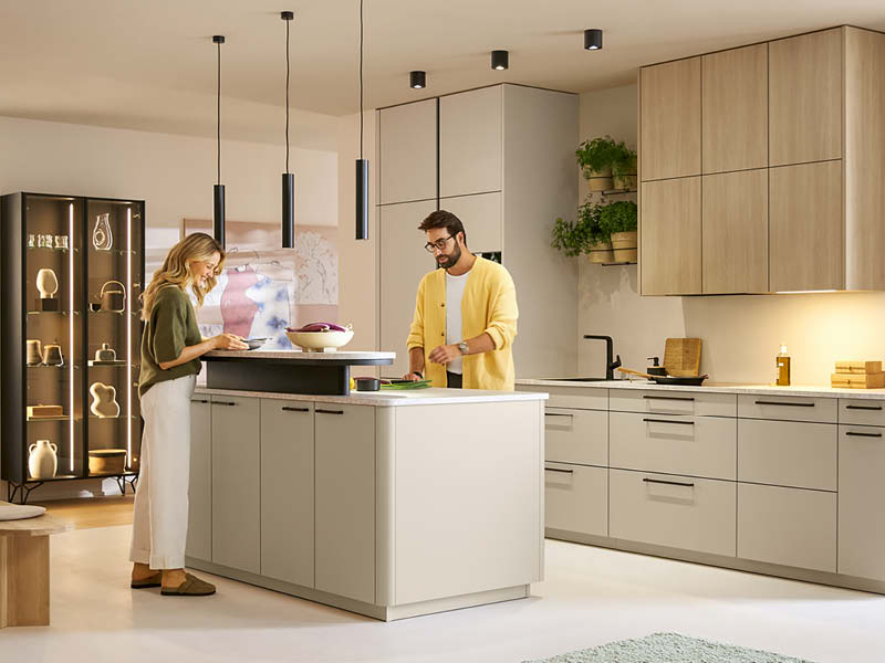 Moderne Schüller Küche mit Kücheninseln in Beige und Holzoptik, integriertem Stauraum und Hängelampen, zwei Personen bereiten gemeinsam Speisen zu