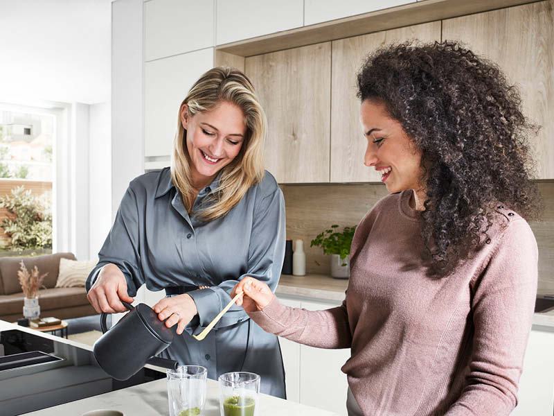 Zwei Frauen bereiten Getränke in moderner Nobilia Küche mit heller Front und Holzrückwand zu, funktionale Arbeitsfläche und wohnliches Design