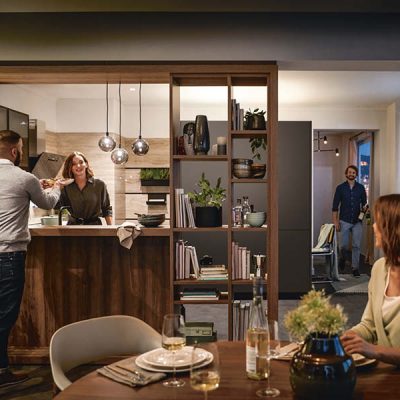 Offene Holzküche mit Gästen beim Kochen im Alltag, warmen Pendelleuchten, Esstisch und wohnlicher Kücheninsel für gesellige Abende.