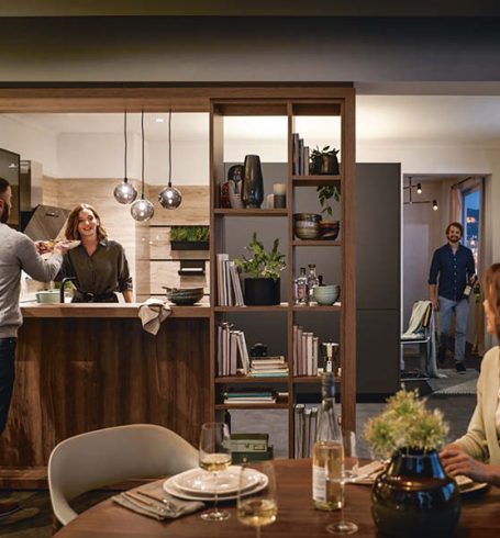 Offene Holzküche mit Gästen beim Kochen im Alltag, warmen Pendelleuchten, Esstisch und wohnlicher Kücheninsel für gesellige Abende.