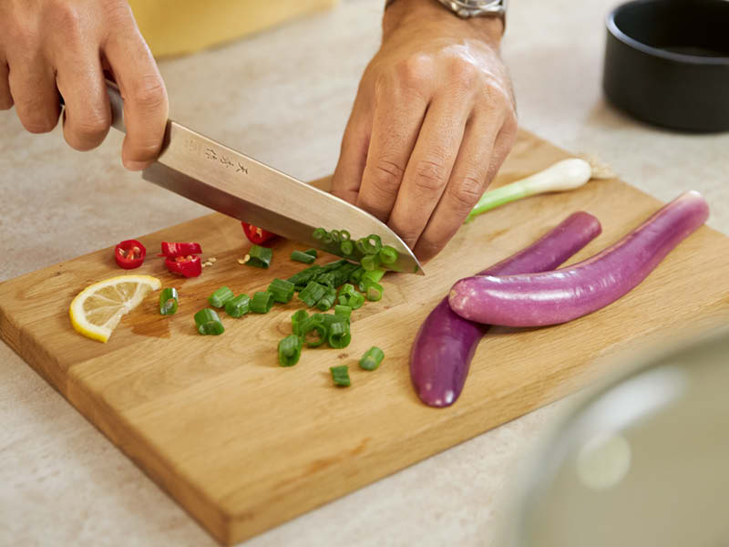 Nahaufnahme vom Schneiden von Frühlingszwiebeln, Chili und Zitrone auf Holzbrett mit Küchenmesser und violetten Auberginen.