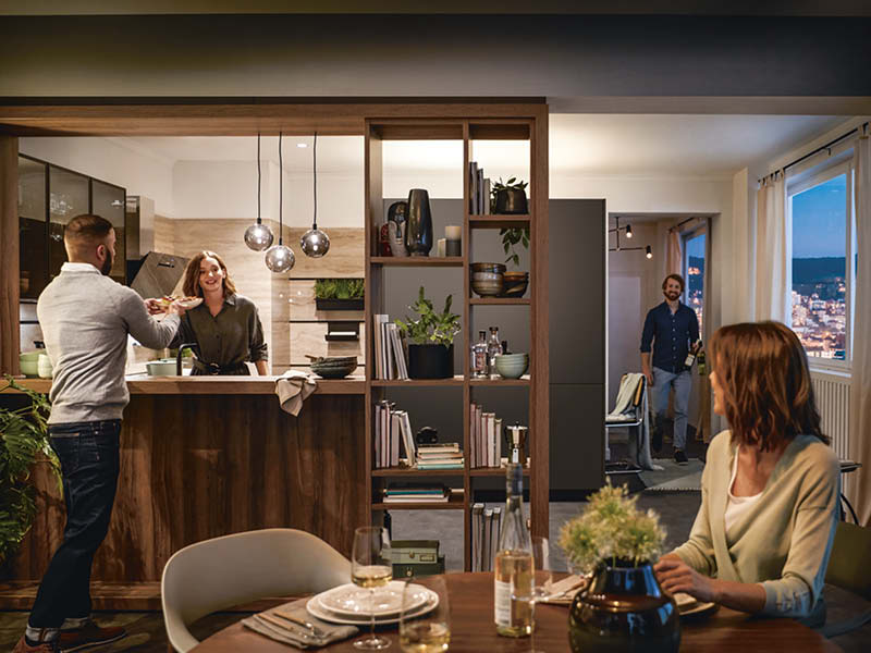 Offene Wohnküche von Schüller mit dunklen Holzfronten, Raumteiler-Regal und Theke, die Kochen, Essen und geselliges Beisammensein stilvoll vereint.