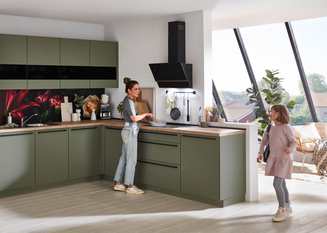 Frau beim Kochen und Kind in einer offenen Küche mit olivgrünen Fronten und Arbeitsplatte in Holzoptik.