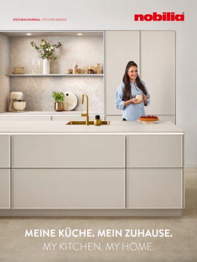 Cover des nobilia Küchenjournals 2027: helle Küche mit Kücheninsel, goldfarbener Armatur und einer Frau mit Tasse.